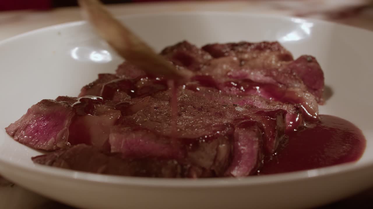 A succulent close-up of perfectly cooked medium-rare beef steak slices, being finished with a glossy red sauce or glaze in a professional kitchen setting