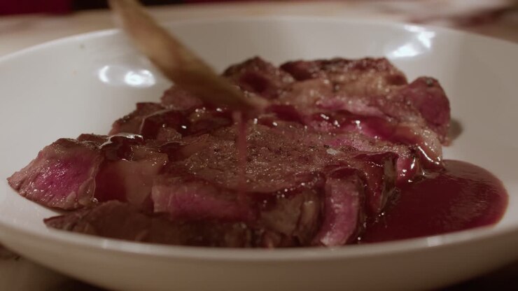 Close-up yang menggugah selera dari irisan steak daging sapi setengah matang yang dimasak dengan sempurna, diselesaikan dengan saus merah mengkilap atau lapisan gula di lingkungan dapur profesional