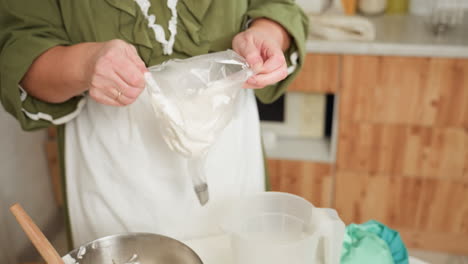 housewife in green top and white apron adjusts transparent piping bag filled with creamy dough while preparing to add more mixture in cozy kitchen with wooden cabinets and countertop baking supplies