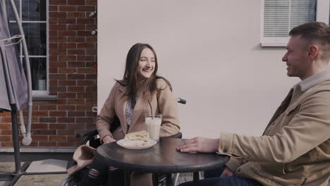 Disabled-Girl-In-Wheelchair-Drinking-Milkshake-And-Talking-With-Her-Male-Friend-In-A-Bar-Terrace