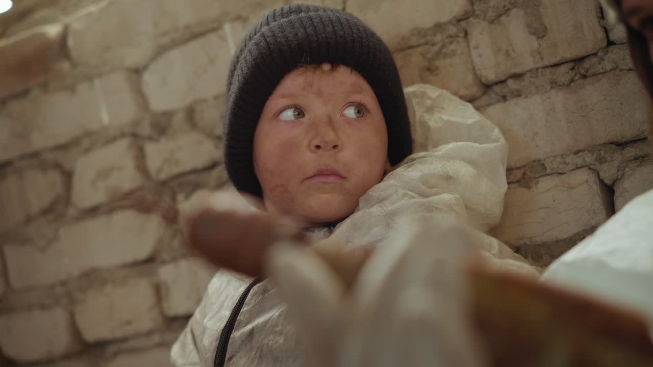 Close up of dirty faced boy in beanie hat sitting against rough brick wall in cold indoor space, wearing worn protective suit, gazing directly with tired expression, possibly in abandoned environment