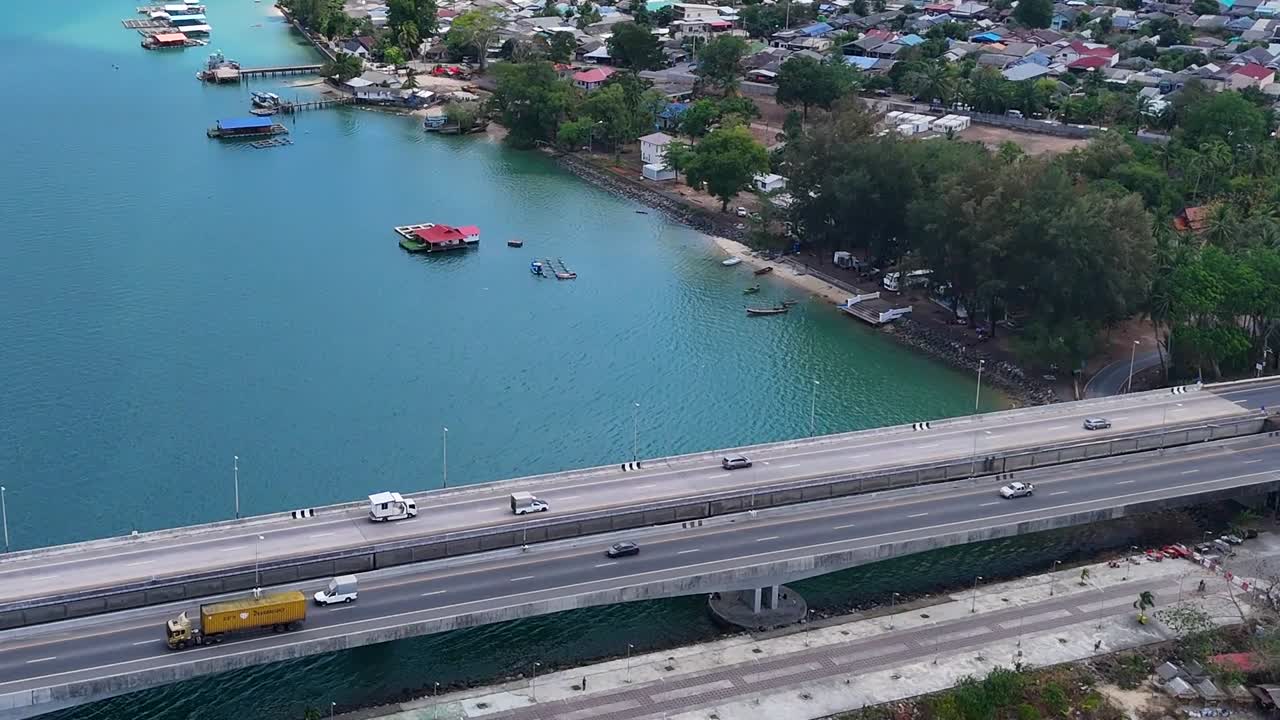 Drone captures a bridge spanning vibrant turquoise waters with nearby greenery and coastal roads.
