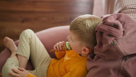 overhead view of young boy lounging comfortably on large pink beanbag while licking mint ice cream cone and gazing toward background where part of stand is visible