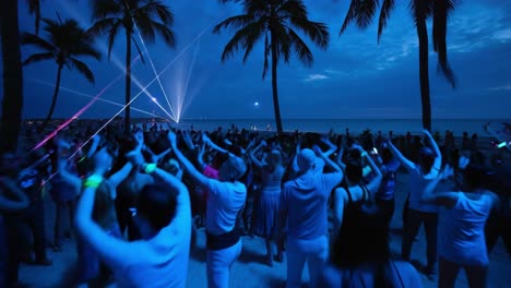 nighttime beach party featuring tourists dancing with glowing sticks, illuminated by stage lights and surrounded by palm trees, creating vibrant tropical celebration atmosphere