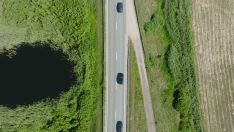 Toma-Aérea-De-Un-Paisaje-Rural,-Camino-Rural-Con-Camiones-Y-Automóviles-En-Movimiento,-Exuberantes-Campos-De-Cultivos-Agrícolas-Verdes,-Día-Soleado-De-Verano,-Amplia-Toma-De-Drones-De-Ojo-De-Pájaro-Descendente