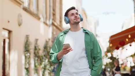 Kaukasischer-Tourist-Mit-Kabellosen-Kopfhörern,-Der-Auf-Dem-Smartphone-Musik-Hört-Und-Durch-Die-Sonnige-Stadtstraße-Geht