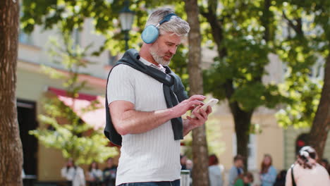 happy mature man in wireless headphones choosing, listening music in smartphone application outdoors