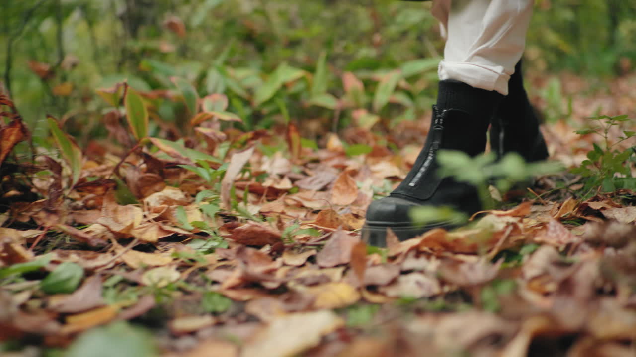 leg view of autumn stroller pushing leaves with black boots on forest path, dry foliage rustling under steps, cozy walk vibe, close ground perspective, soft bokeh, tranquil nature mood