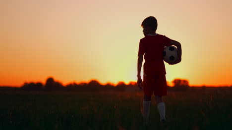 Der-Junge-Geht-Bei-Sonnenuntergang-Mit-Einem-Fußball-Auf-Das-Feld,-Blickt-In-Die-Ferne-Und-Träumt-Davon,-Ein-Erfolgreicher-Fußballspieler-Zu-Werden.-Beobachten-Sie-Den-Sonnenuntergang-Auf-Dem-Feld