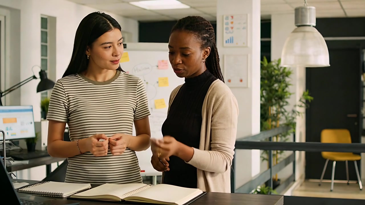 Reviewing two women in office striped tee and cardi gesturing, checking pen, open notebook starting