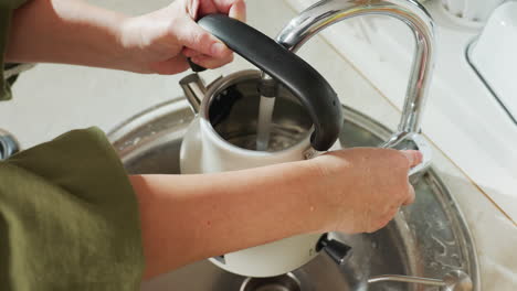 overhead view of woman filling electric kettle with water from kitchen tap, carefully turning off faucet when full, with water droplets on hands and reflective stainless steel sink visible beneath