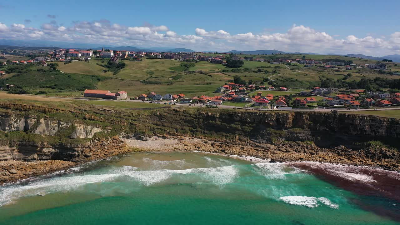 fotografía aérea en verano en la costa del mar hacia el acantilado en el que corre una carretera con varias casas típicas enmarcadas en prados verdes con el cielo azul en el fondo y algunas nubes en cantabria, españa