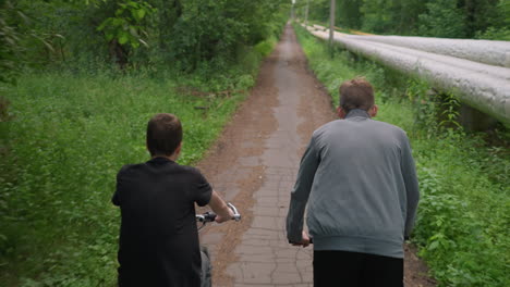 two friends riding bicycles along a paved untarred path surrounded by lush greenery and trees, one stands while cycling, and a pipe lies along the side of the path