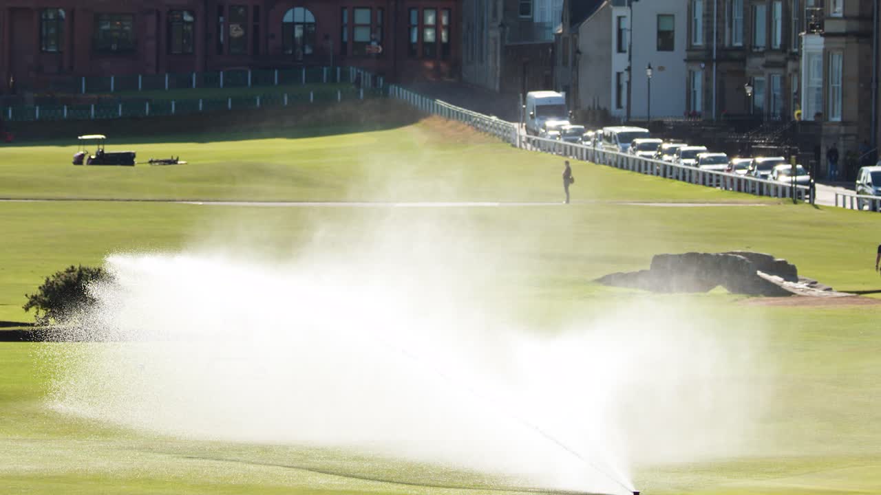 Rotating sprinkler sprays water across sunlit golf course, distant buildings, and lone figure visible