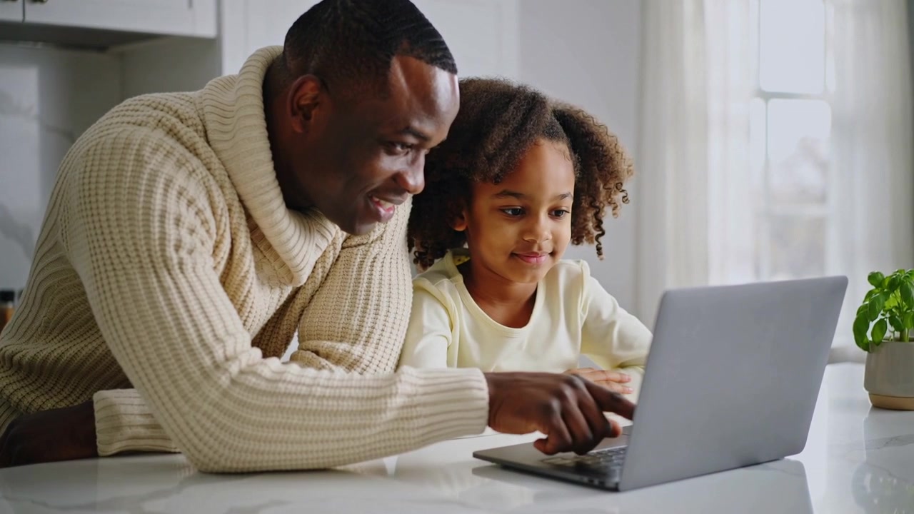 Leaning father teaching daughter, pointing at laptop trackpad on kitchen counter, in turtleneck
