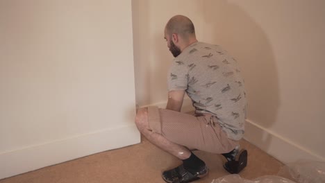 Joven-Adulto-Caucásico-De-Rodillas-Pintando-La-Pared-Blanca-De-Una-Habitación-Vacía-En-Casa-Con-Rodillo-Cambridge,-Inglaterra