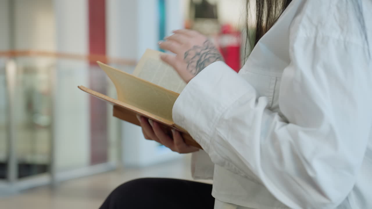 Side view of lady seated holding brown diary with visible hand tattoo, facing forward in well-lit shopping mall interior, blurred figures and clothing store in background