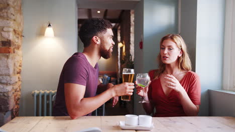 mixed race young adult couple sitting at table in a pub holding drinks and talking, side view