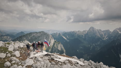 Drohnenaufnahme-Von-Der-Spitze-Des-Berges-Rombon,-Eine-Gruppe-Von-Wanderern-Mit-Einer-Slowenischen-Flagge,-Im-Hintergrund-Ein-Tal-Und-Berge,-Der-Himmel-Ist-Mit-Wolken-Bedeckt