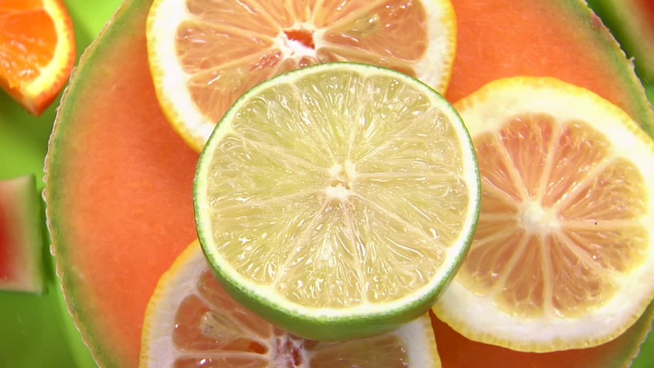 Yeşil bir tabakta dilimlenmiş misket limonu, limon, portakal ve karpuz içeren canlı bir meyve düzenlemesinin yakın çekim görüntüsü. Simetrik tasarım, meyvelerin tazeliğini ve çeşitliliğini ön plana çıkarıyor.
