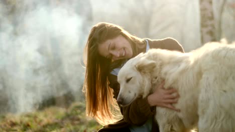 smiling happy woman gently stroking white dog around fire place.real friends people outdoor camping tent vacation in autumn trip.fall sunny day in nature, animal friendship. 4k video