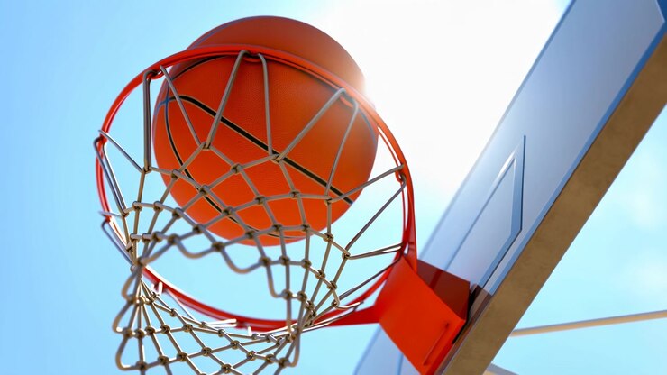 Vista ravvicinata dal basso di un pallone da basket arancione che cade attraverso un canestro rosso lucido e una rete di nylon intrecciata accanto a un tabellone traslucido, con luce solare intensa, cielo blu e una forte energia da campo all'aperto.
