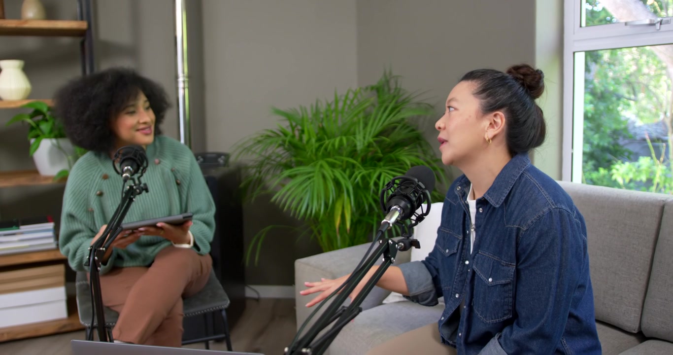 Diverse women hosts starting podcast in studio holding tablet, guest speaking, fixing mic, smiling