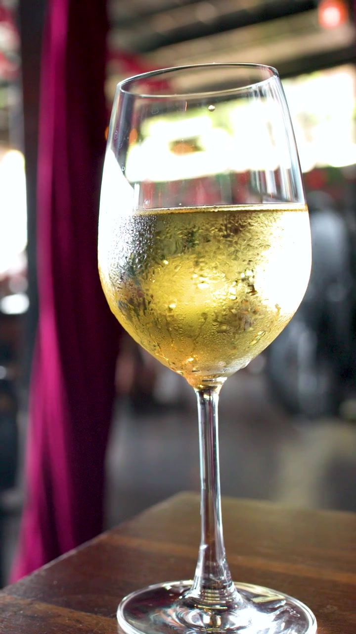 Un bicchiere di vino bianco coperto di condensa è posato su un tavolo di legno in un bar all'aperto.