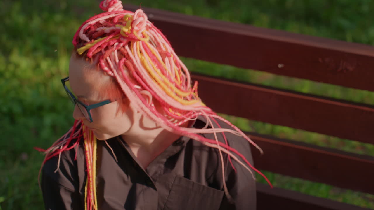 다채로운 머리 움직임: 밝은 여성분이 자연광 아래에서 생기 넘치는 분홍색 머리카락을 활기차게 가지고 놀아요. 활발한 머리카락이 춤추고 다채로운 땋은 머리가 야외에서 흔들리는 역동적인 장면이에요.