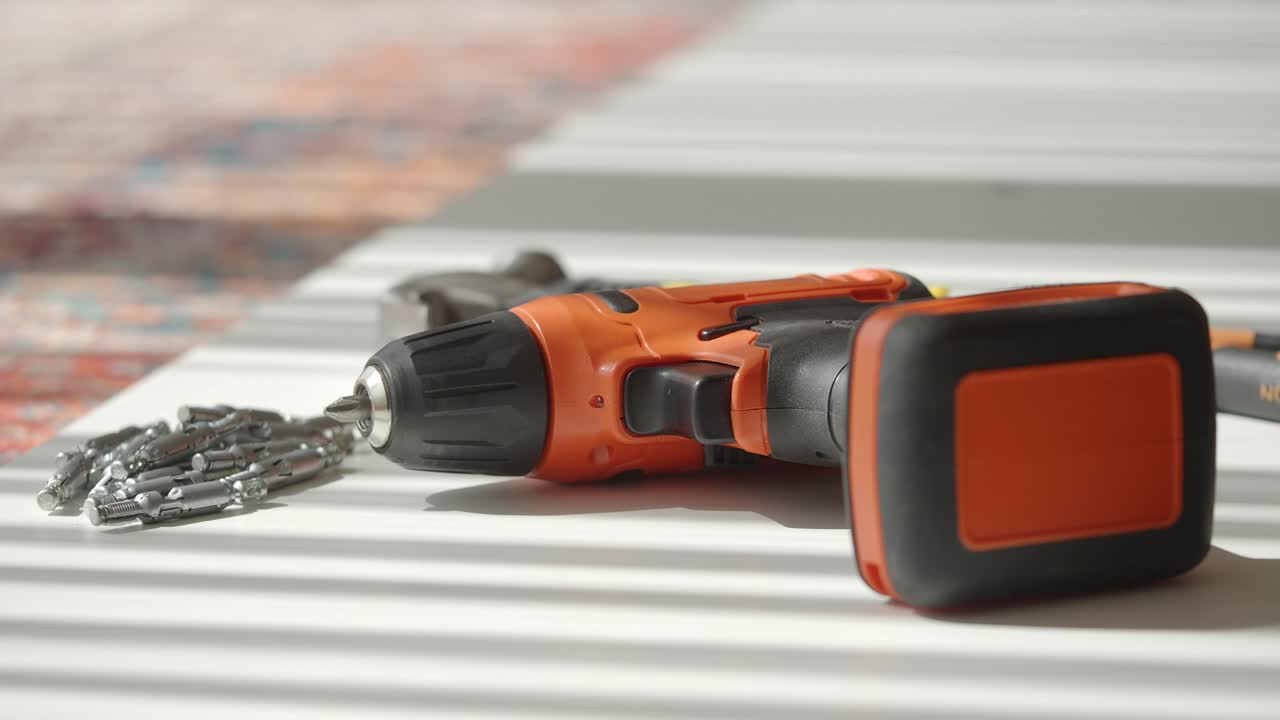 A collection of DIY and construction tools, including a power drill, drill bits, hammer, and pliers, with a person's hand interacting with the drill