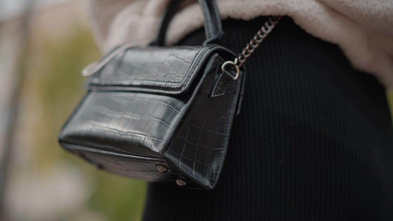 Vista ravvicinata di una piccola borsa in pelle nera sospesa a una catena di metallo e che oscilla accanto a un capo di abbigliamento in maglia a coste scura durante una passeggiata all'aperto, con luce diurna morbida e diffusa, profondità di campo ridotta e un'atmosfera calma.