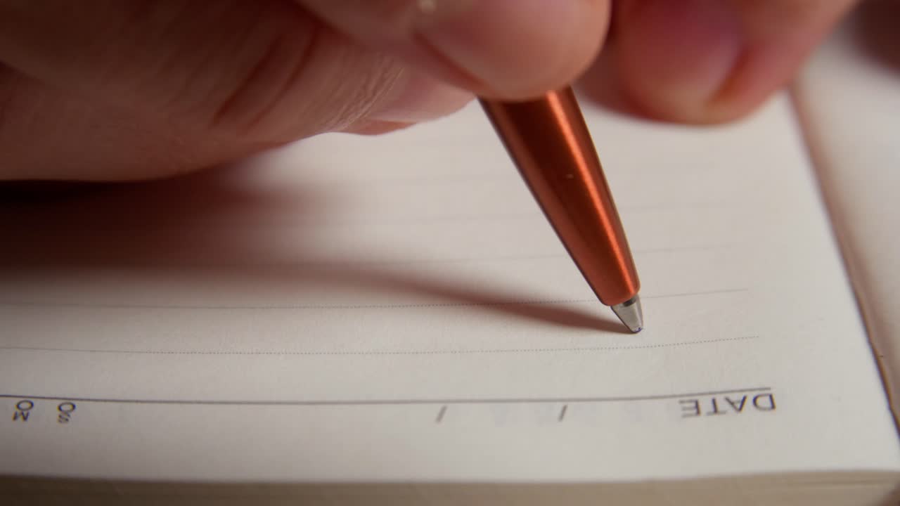 Man sits with notebook and pen at an indoor table. He is struggling with what to write. Lists goals to achieve. Close in shot. Clip 3