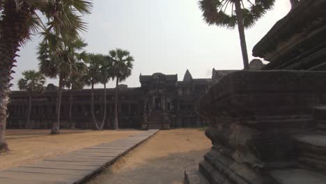 Dolly-shot-along-the-steps-of-a-temple-in-Cambodia's-Angkor-Wat,-showing-parmtrees-and-ancient-temples,-bright-blue-sky-day