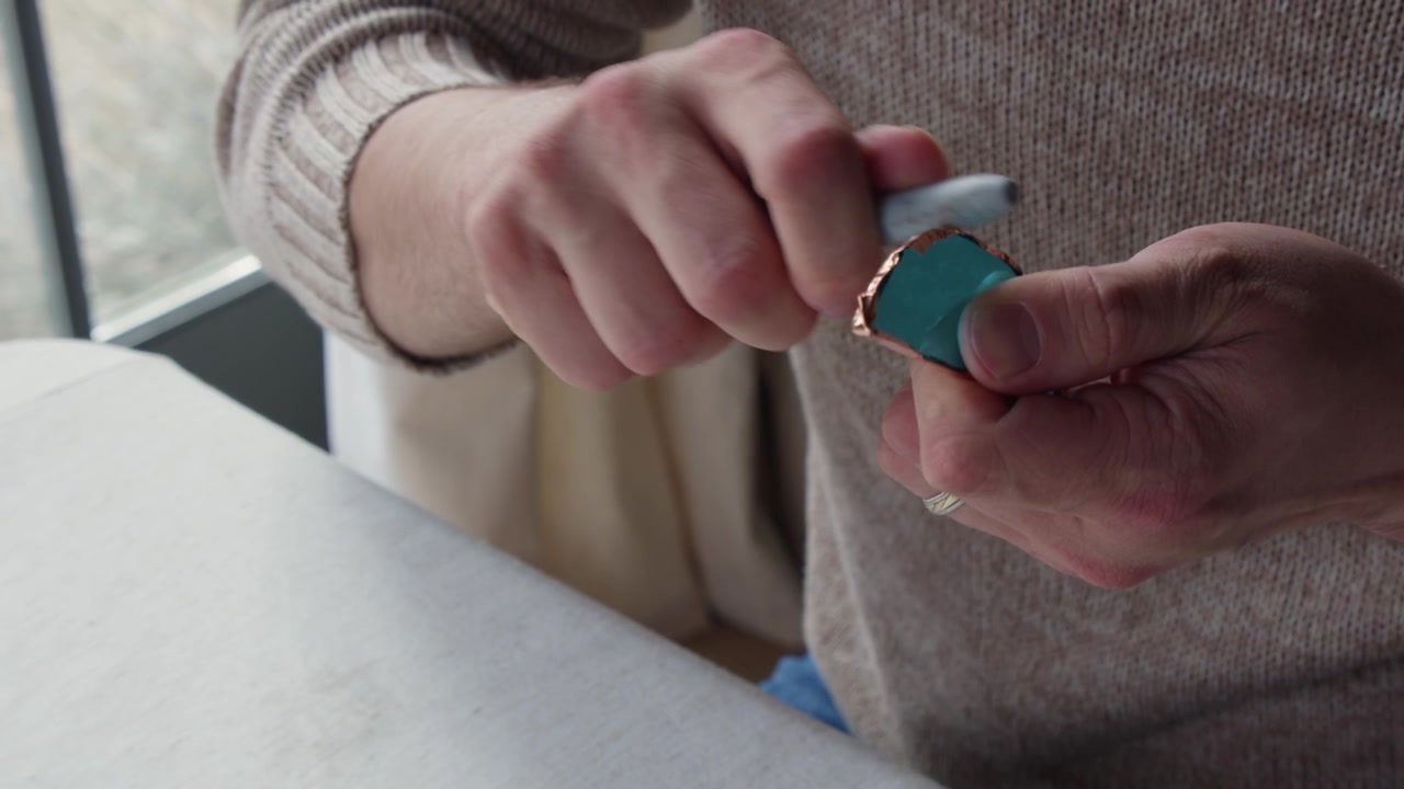 A male art student wearing a beige sweater uses the back of a pen to mold copper tape onto a piece of cut, blue stained glass.