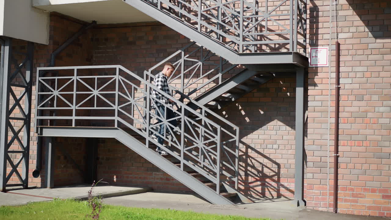 side view of student descending metal staircase pauses placing cigarette between lips sparks lighter tip as green grass sways in wind against brick and metal backdrop under warm sunlight