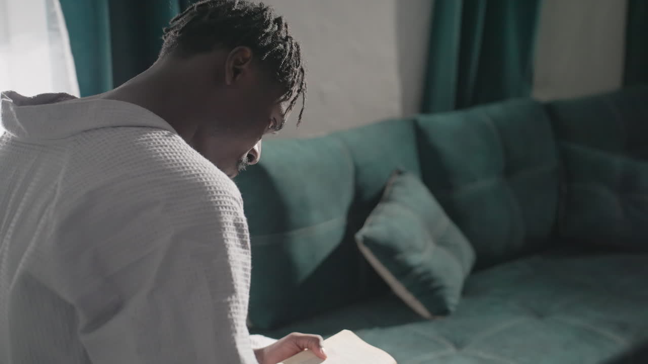 Guy in bathrobe sitting on bed, deeply engrossed in reading book. Natural light pours in from window, creating peaceful, cozy ambiance. Tranquil, intimate moment in a quiet, comfortable room