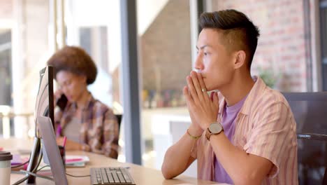 Nachdenklicher-Asiatischer-Mann,-Der-Den-Computer-Am-Schreibtisch-In-Einem-Ungezwungenen-Büro-Benutzt,-Mit-Verschiedenen-Kollegen,-Zeitlupe