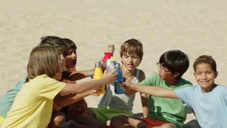 excited boys sitting on beach and clinking with water bottles