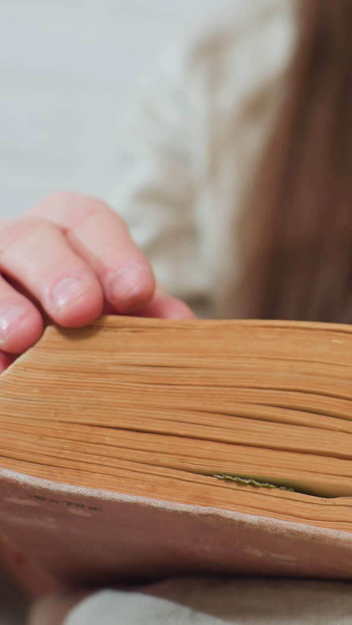 두꺼운 책 페이지를 넘기는 손, 텍스트와 노트를 찾는 백인 학생, 클로즈업 스티치 바인딩, 여백 흔적, 소파 위의 자연광, 집중적인 학습 의식과 조용한 발견