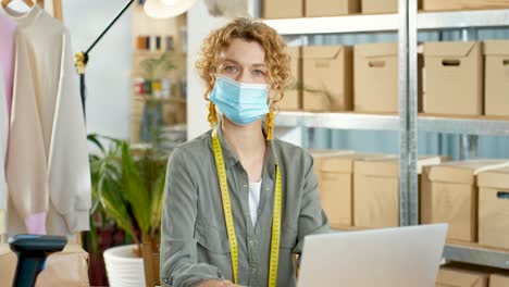 Young-caucasian-designer-woman-in-facial-mask-and-typing-on-laptop-at-office-in-clothing-shop