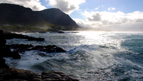 Wave-reflecting-beautiful-morning-light-as-it-rolls-past-onto-rocky-coastline,-with-mountains-in-background