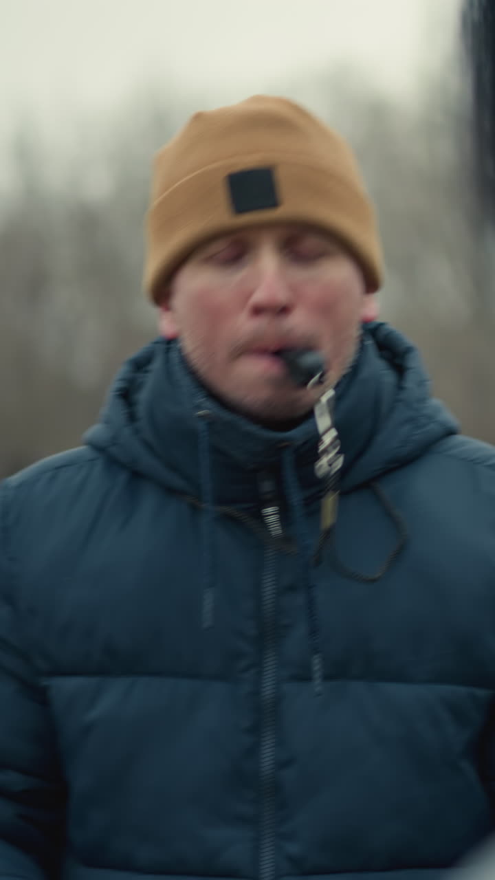 A coach holding a soccer ball in a field with a whistle in his mouth, playfully hits someone on the head with the ball, the background features blurred trees and a goal post