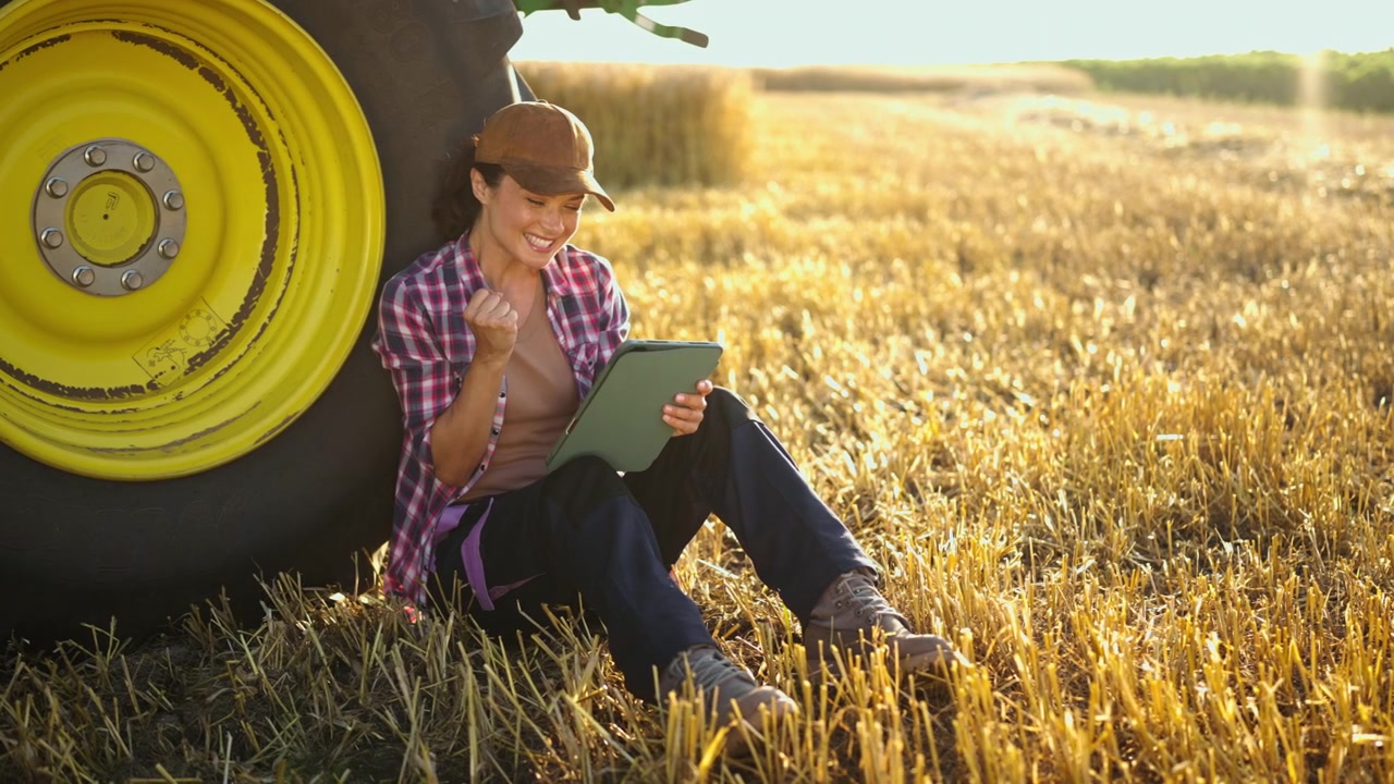 Agricoltrice che Usa un Tablet Seduta accanto al Trattore in un Campo Raccolto