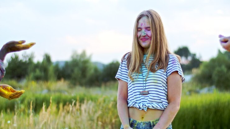 Young Pretty Girl Standing In The Field And Laughing While Her Friends Tossing Colorful Paints In Powder During Holi Fest