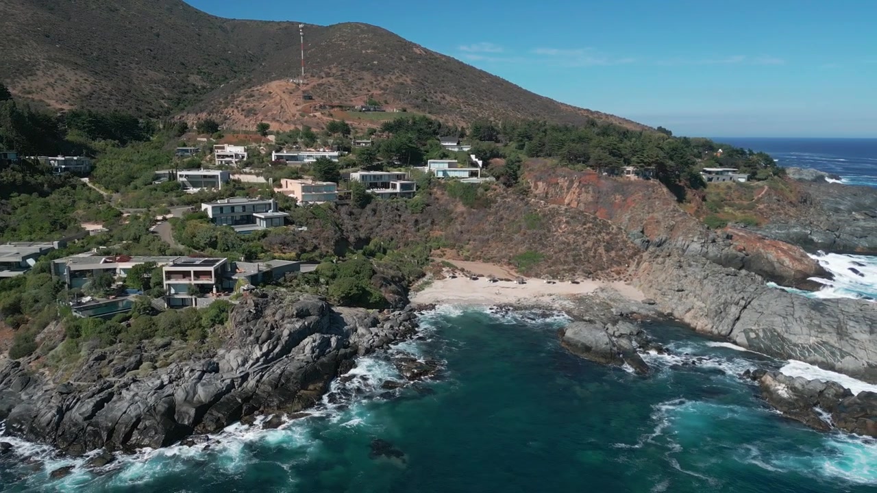 Vista panorâmica sobre villas exclusivas no topo do penhasco com enseada de areia isolada, promontório rochoso acidentado e colina de vegetação rasteira seca ao fundo. Punta Pite, Papudo, Chile