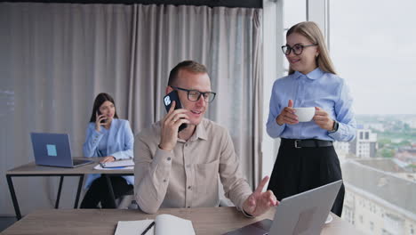 business conversation on the phone. man speaking with clients on the phone and woman standing beside with a cup in hand points at the screen. female coworker at backdrop.