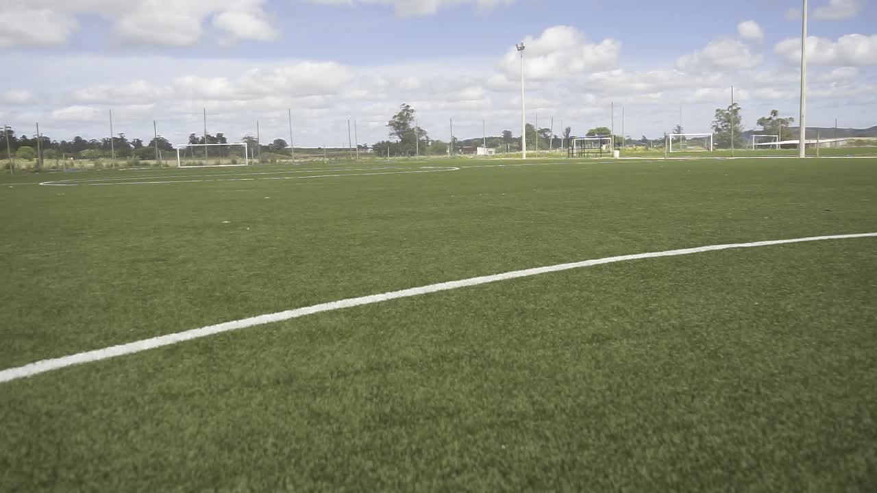 Camera panning across lush green soccer field, revealing crisp white boundary lines and empty goal areas under bright summer sky with soft clouds