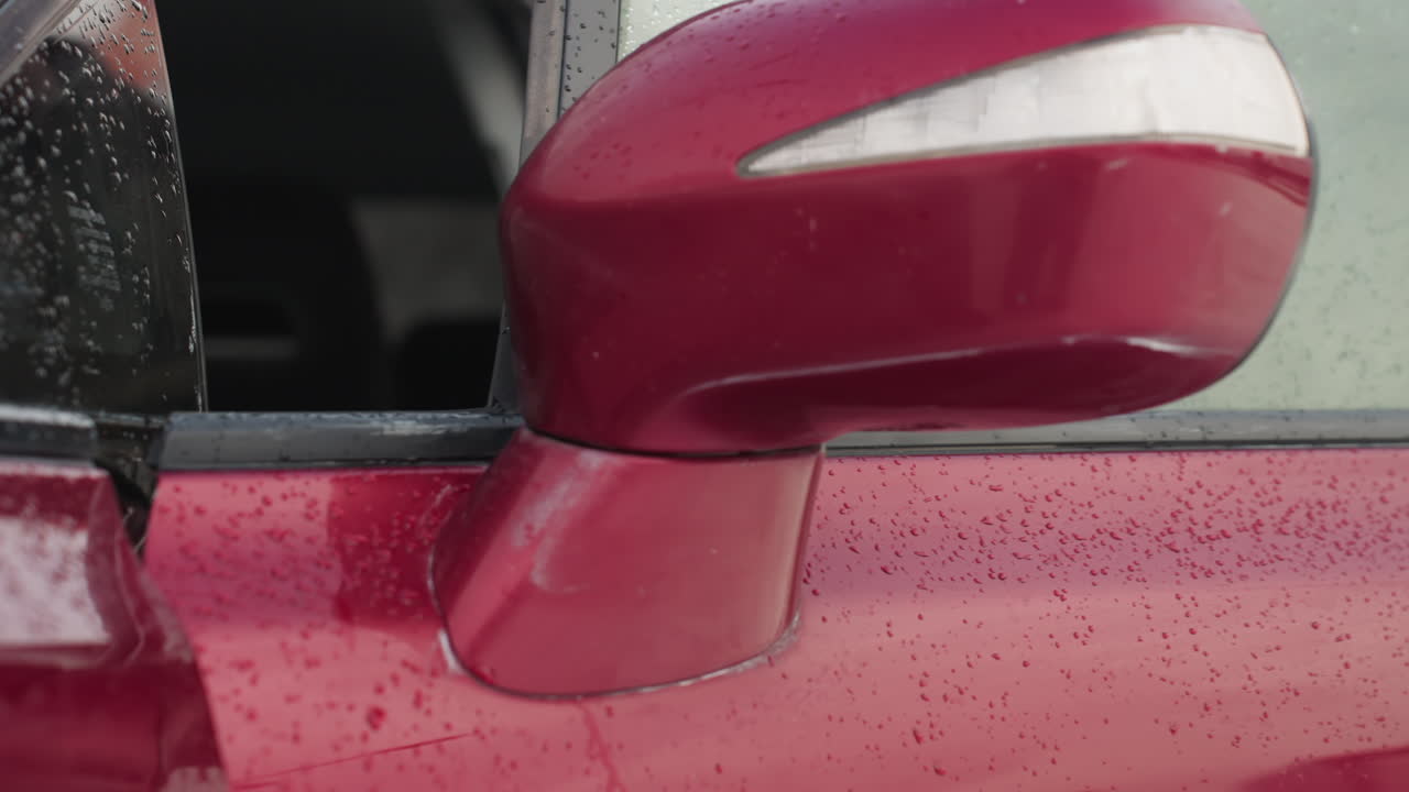 Close up of red car side mirror with water droplets on surface during cold winter day, soft background with snow-covered trees and blurred figure opening car door