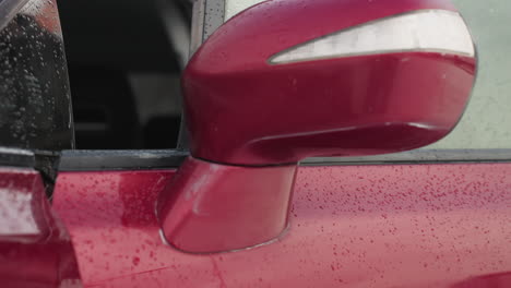 close up of red car side mirror with water droplets on surface during cold winter day, soft background with snow-covered trees and blurred figure opening car door