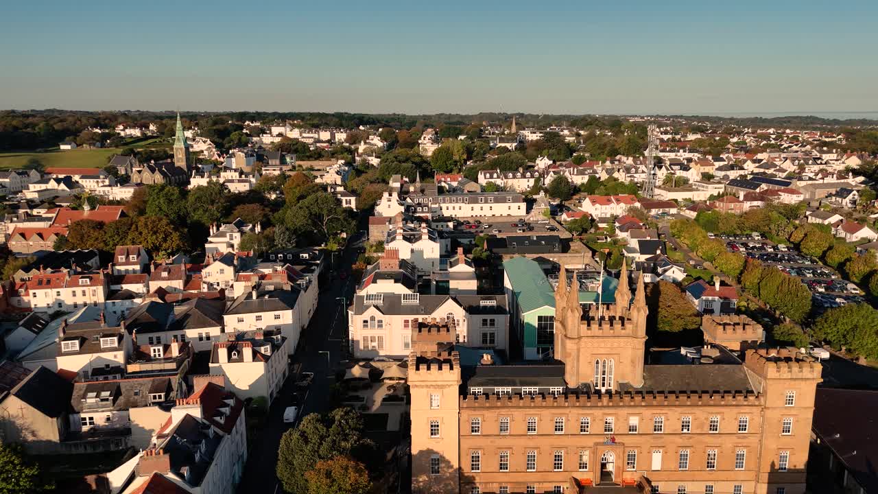 Långsam drönarfilmning på låg höjd över Saint Peter Port, Guernsey, över Elizabeth College och stadens takåsar med utsikt över stadssilhuetten och hela ön en molnfri sommardag i starkt solsken.